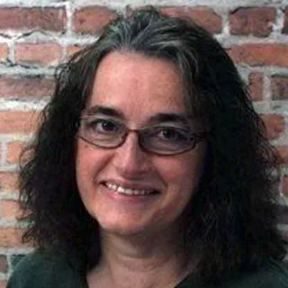 Lächelnde Frau mit dunklen Haaren und Brille, Ziegelstein-Hintergrund.