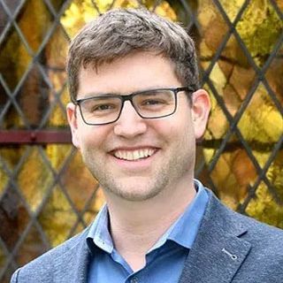 Lächelnder Mann mit Brille vor Buntglasfenster.