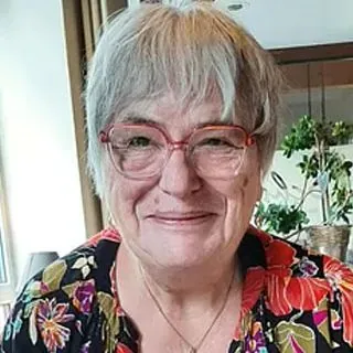 Lächelnde Frau mit kurzen grauen Haaren, Brille, geblümtem Oberteil.