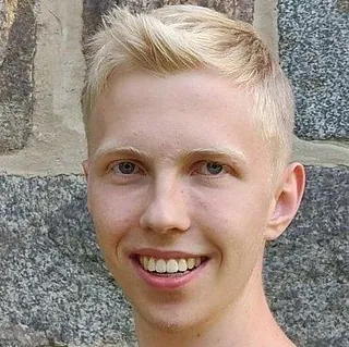 Lächelnder junger Mann mit blonden Haaren vor Steinmauer.