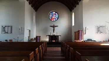 Innenansicht einer Kirche mit Bänken, Altar, Kruzifix und Buntglasfenster.