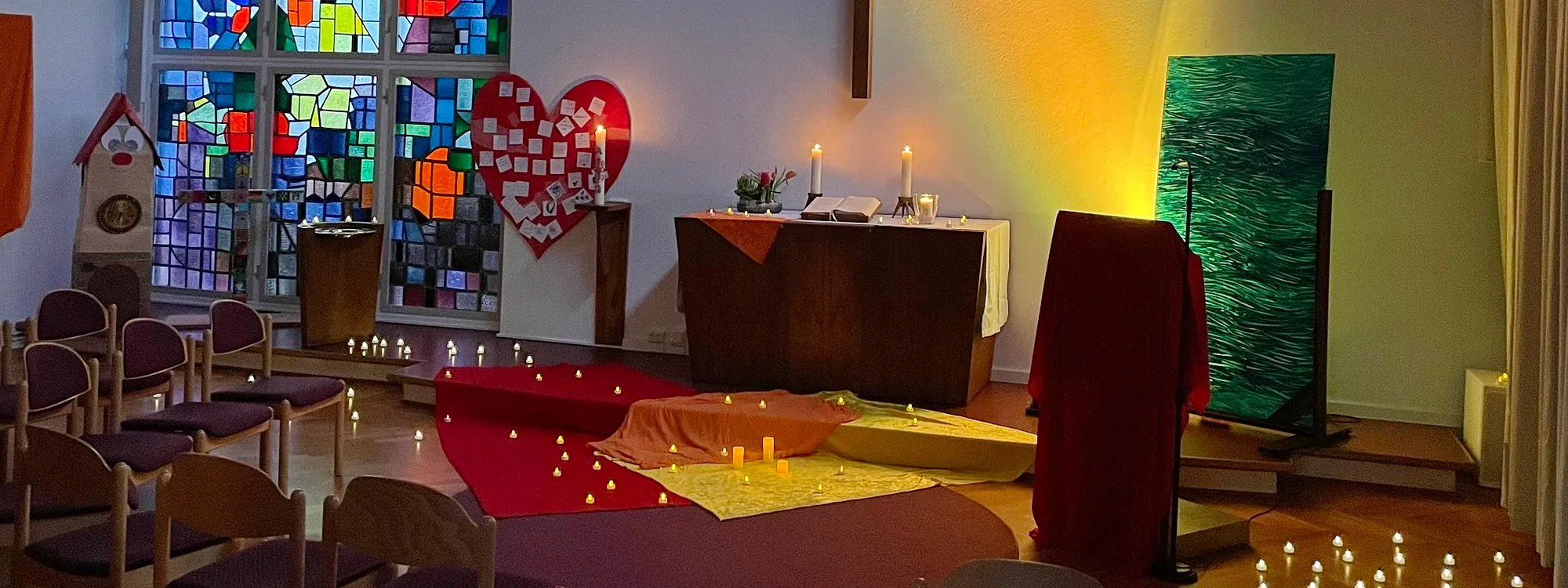 Kirchraum mit Altar, buntem Fester, Kerzen auf den Boden und rote und gelbe Tücher