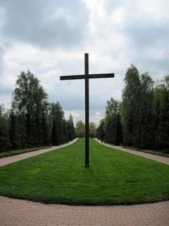 Freistehendes Kreuz auf Rasenfläche mit Bäumen, bewölkter Himmel.