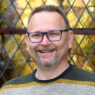 Lächelnder Mann mit Bart und Brille, Buntglas-Hintergrund.